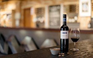 A bottle of wine sits atop a table with a glass next to it in a winery