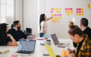 Woman stands at head of meeting table pointing to a group of post-it notes on the wall